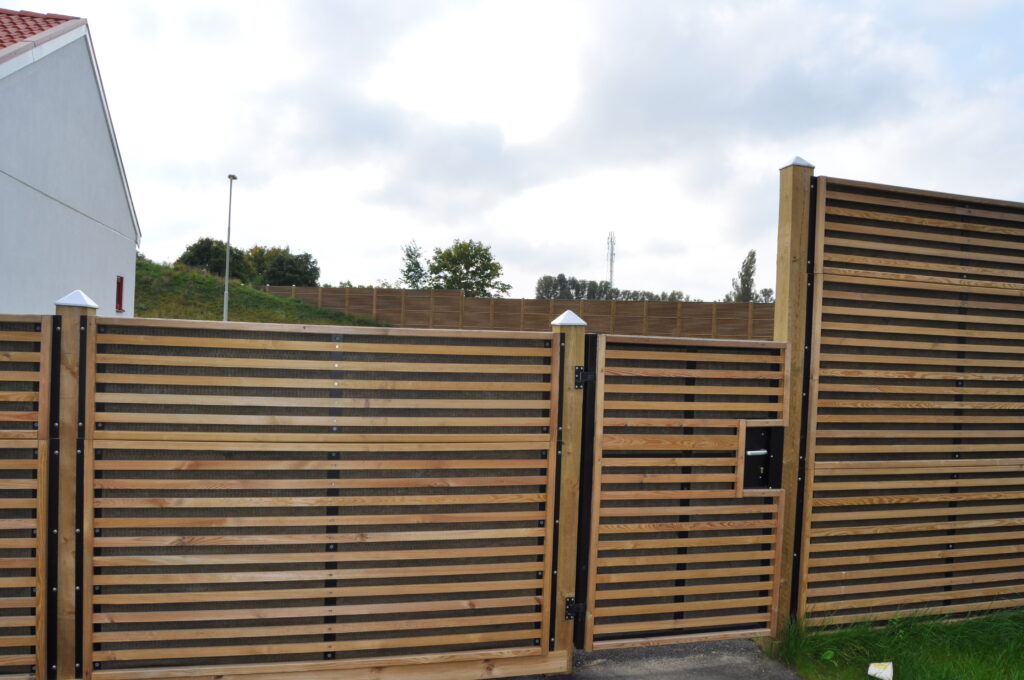 Tall acoustic fence in a domestic garden. The fence is clad in wood batons and encloses a garden. The acoustic fence also has an acoustic gate. 