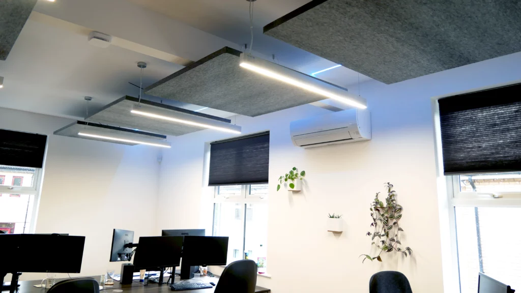 Acoustic baffles hanging from a ceiling in an office. There are computers, desks, office chairs, windows and plants. The office is contemporary.